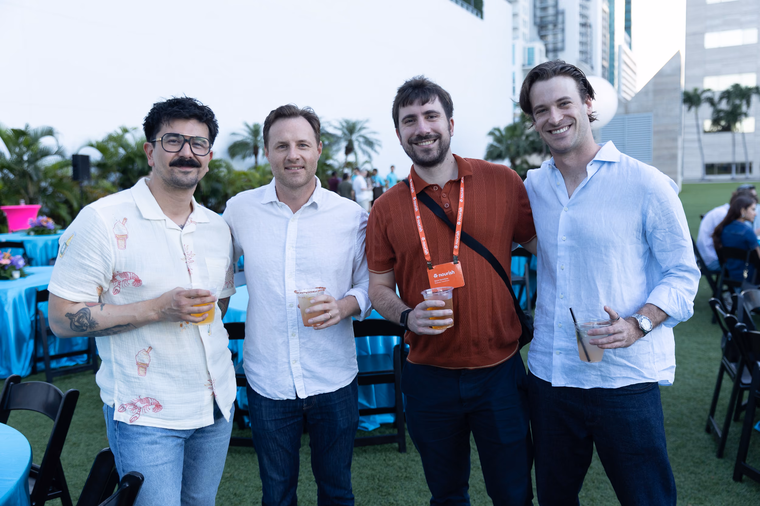 Four men standing outdoors with drinks at a casual event, smiling at the camera.