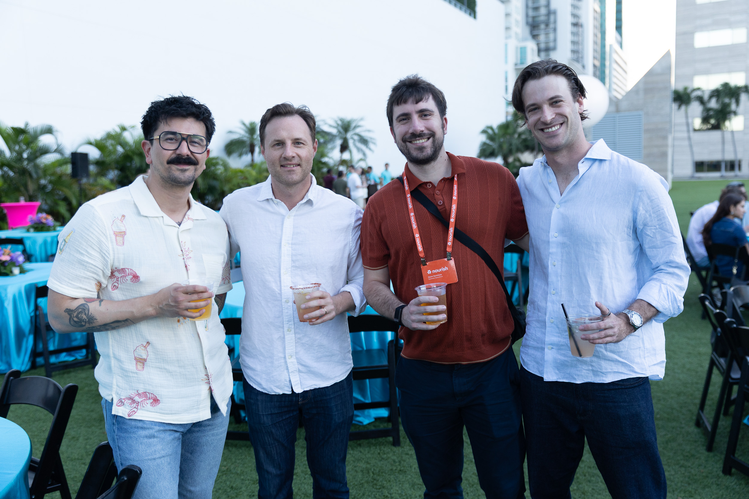 Four men standing outdoors with drinks at a casual event, smiling at the camera.