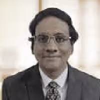 Portrait of a man with glasses wearing a suit and patterned tie, smiling slightly against a blurred indoor background.
