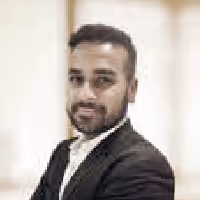Professional man in a black suit with folded arms smiling against a blurred light background.