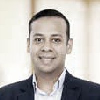 Smiling man with short hair wearing a dark blazer and light shirt against a light background.