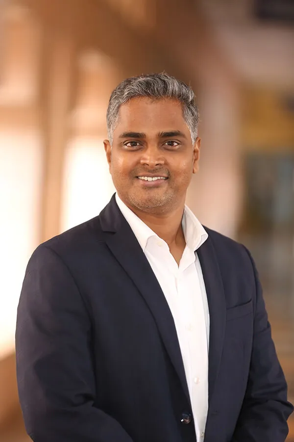 Smiling man with grey hair wearing white shirt and dark blazer on blurred warm background.