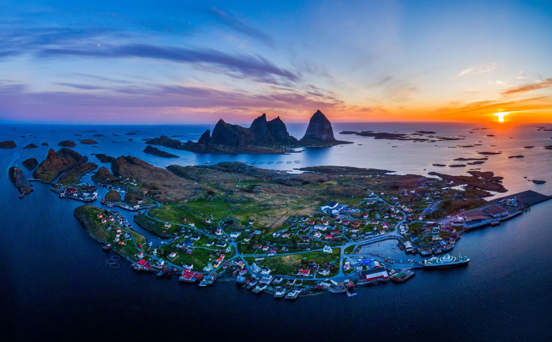 Panoramisk luftbilde av et kystsamfunn med dramatiske fjellformasjoner i bakgrunnen ved solnedgang