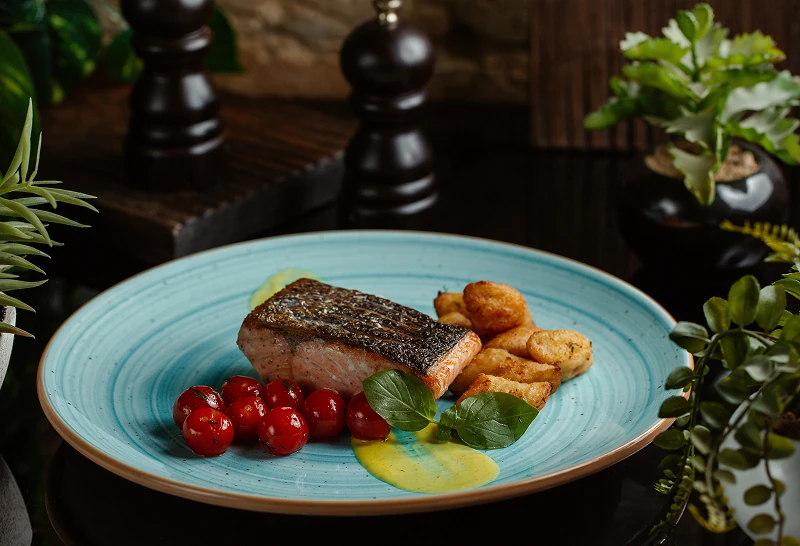 Grilled salmon fillet served with cherry tomatoes, fried potato bites, and a yellow sauce on a blue plate.