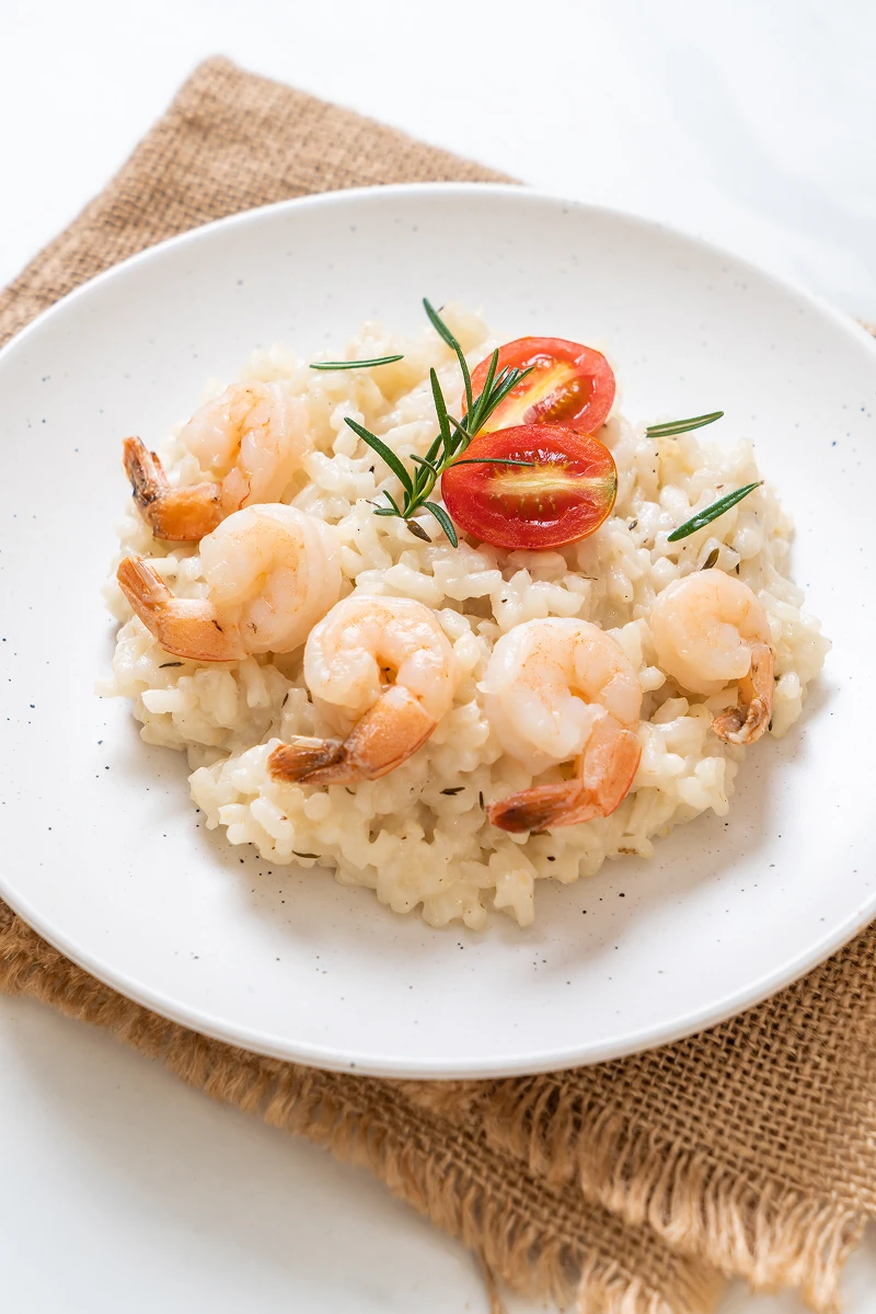 White plate of creamy risotto garnished with cooked shrimp, cherry tomato halves, and rosemary sprigs on a brown textured cloth.