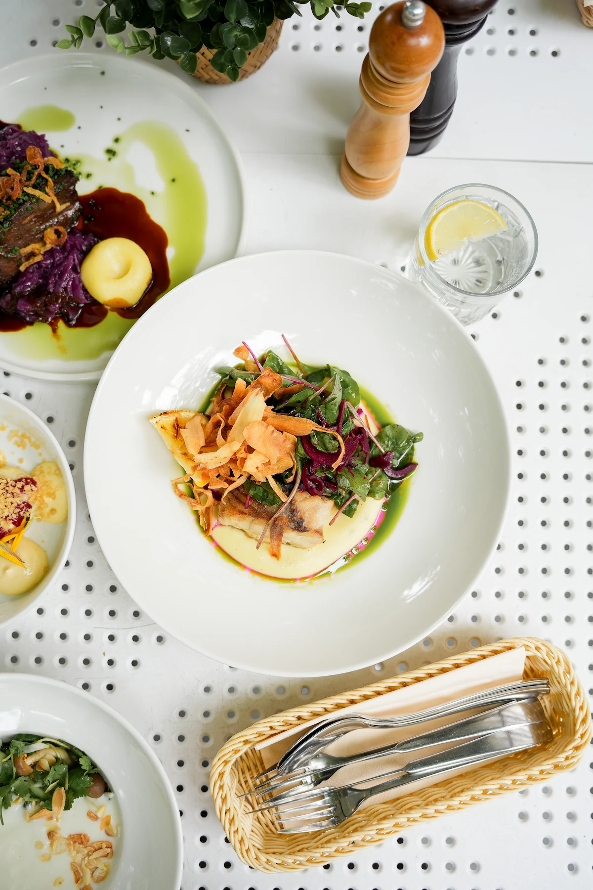 White bowl with a gourmet dish of fish topped with greens and crispy garnish, surrounded by plates of food, a glass of water with lemon, pepper mills, and cutlery on a white perforated table.