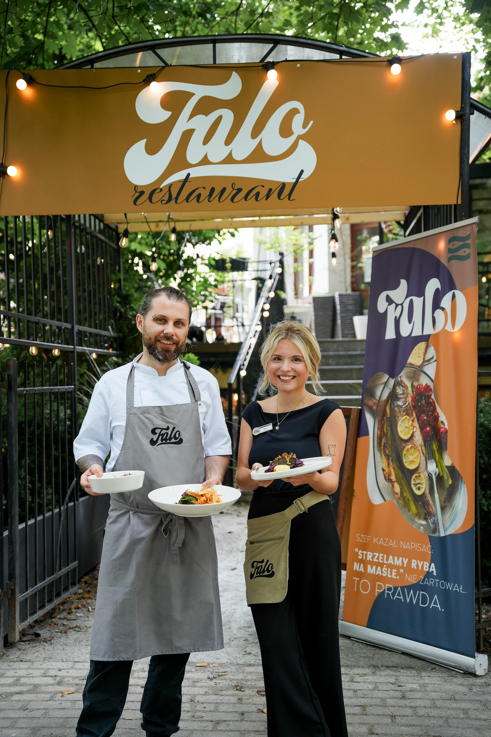Smiling male chef and female server holding plated dishes in front of Falo restaurant yellow sign and banner.