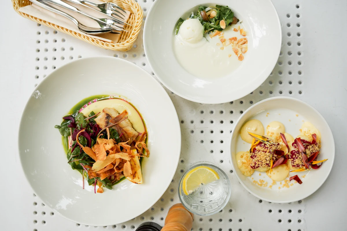 Top view of three gourmet dishes on white plates, a glass of lemon water, and a basket of cutlery on a white perforated table.