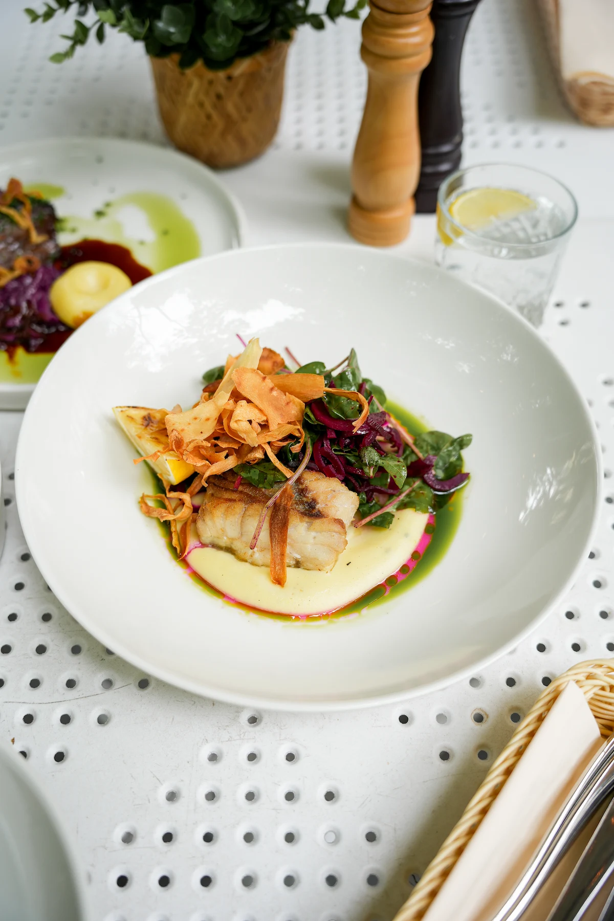 Plated cooked fish fillet served with leafy greens, crispy root vegetable chips, and creamy sauce on a white plate.