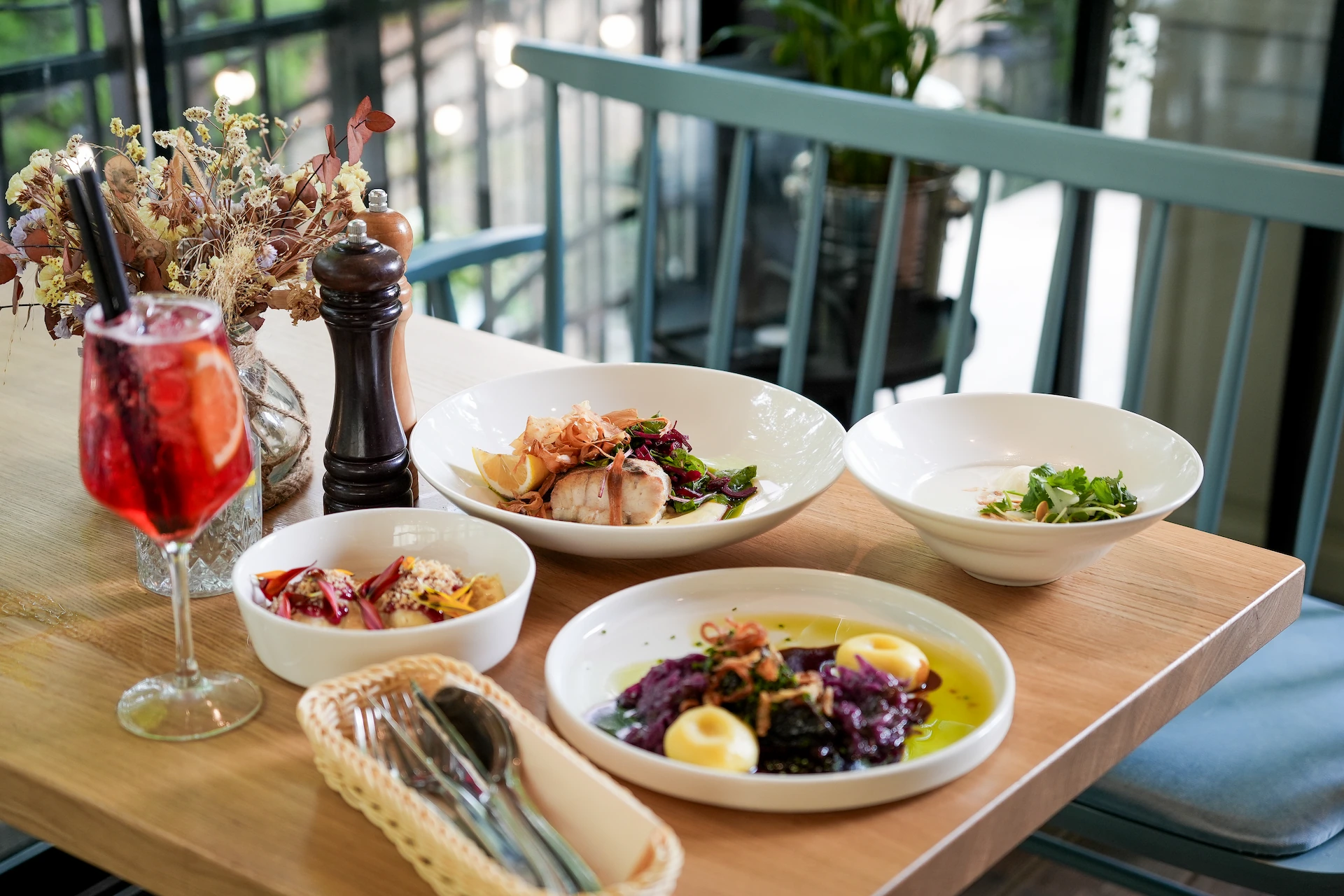 Table with various plated gourmet dishes, a red beverage in a glass with two black straws, dried floral arrangement, and cutlery in a basket.