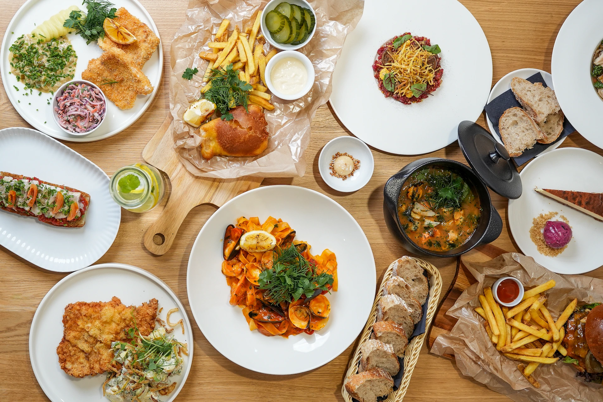 Top-down view of a wooden table filled with various dishes including seafood pasta, fried fish with sides, French fries with tartar and pickles, bread slices, soup, a burger with fries, and a dessert plate.