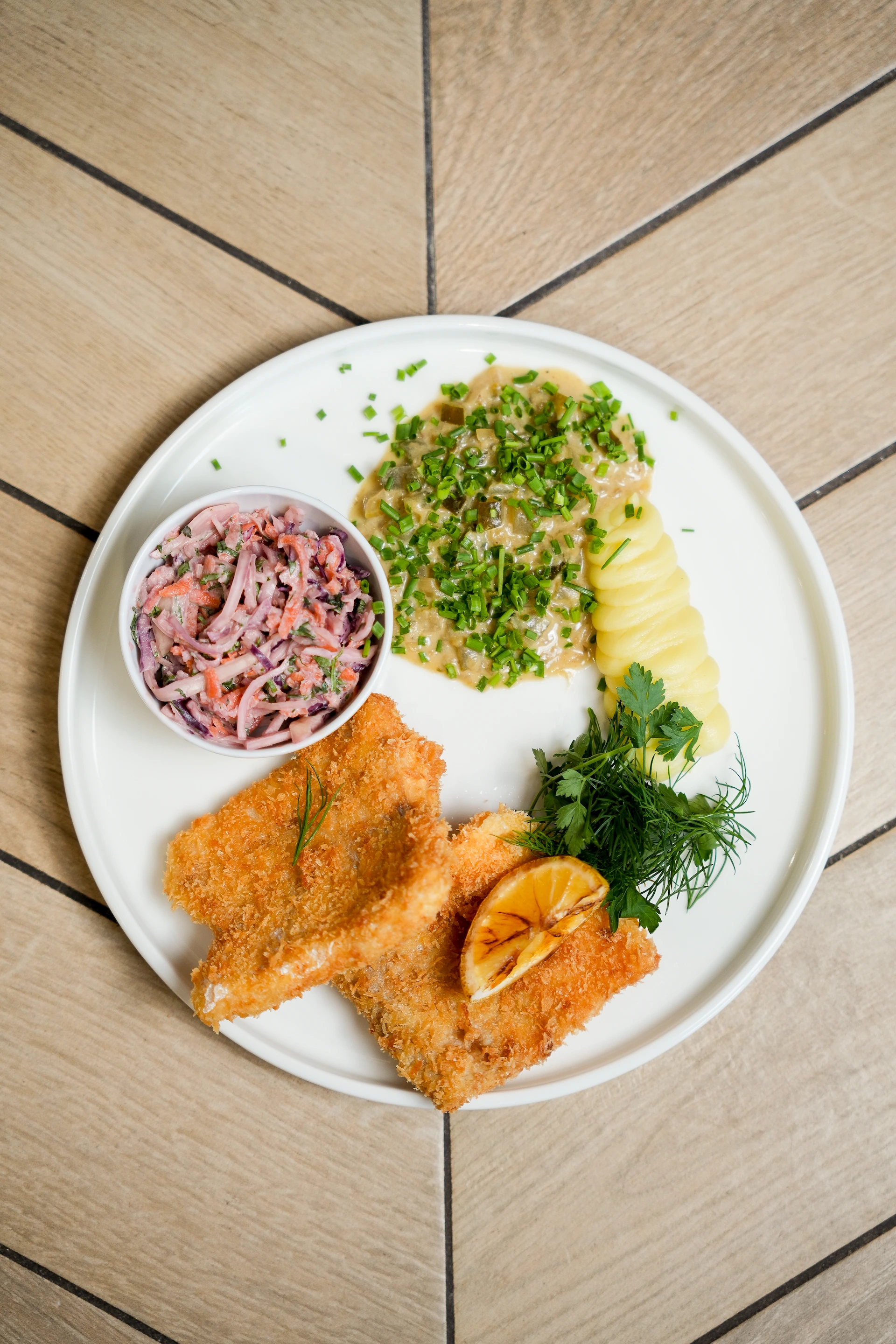Plate with two breaded fried fish fillets garnished with grilled lemon and herbs, creamy potato slices with green garnish, sauce topped with chopped chives, and a small bowl of coleslaw.