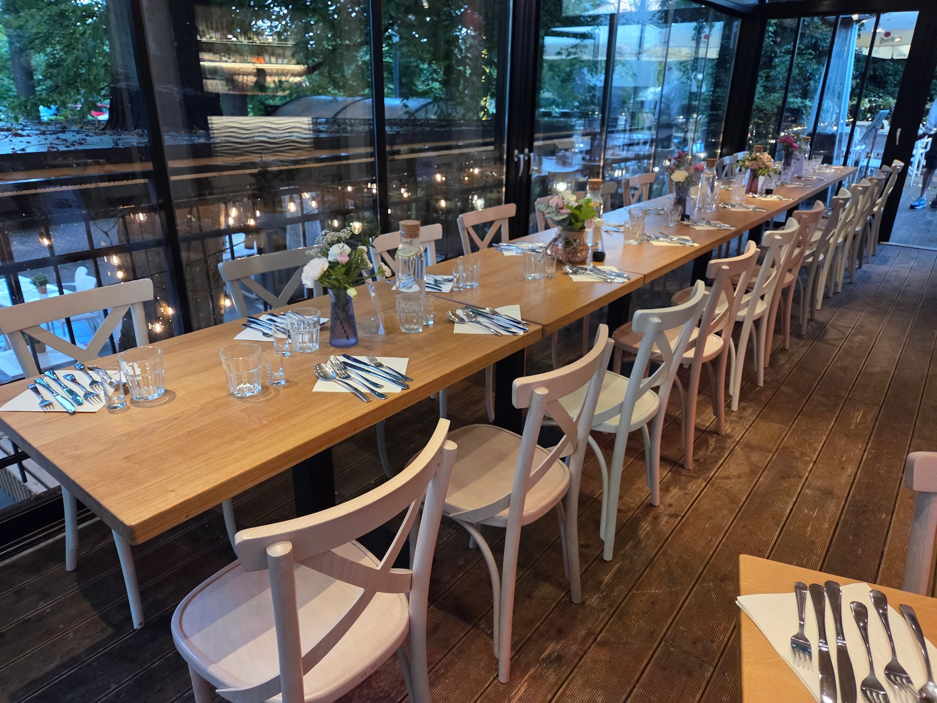 Long wooden table with white wooden chairs set for a meal, featuring cutlery, glasses, and small flower arrangements inside a glass-walled restaurant.