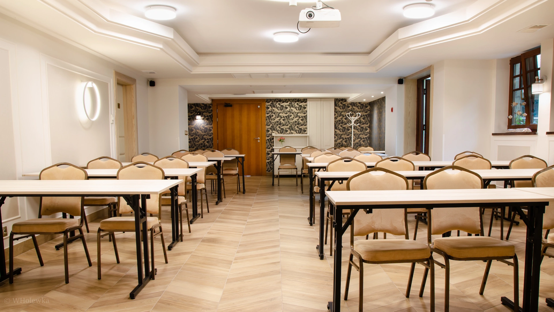 Bright conference room with rows of beige chairs and white tables arranged in neat lines facing a wooden door.