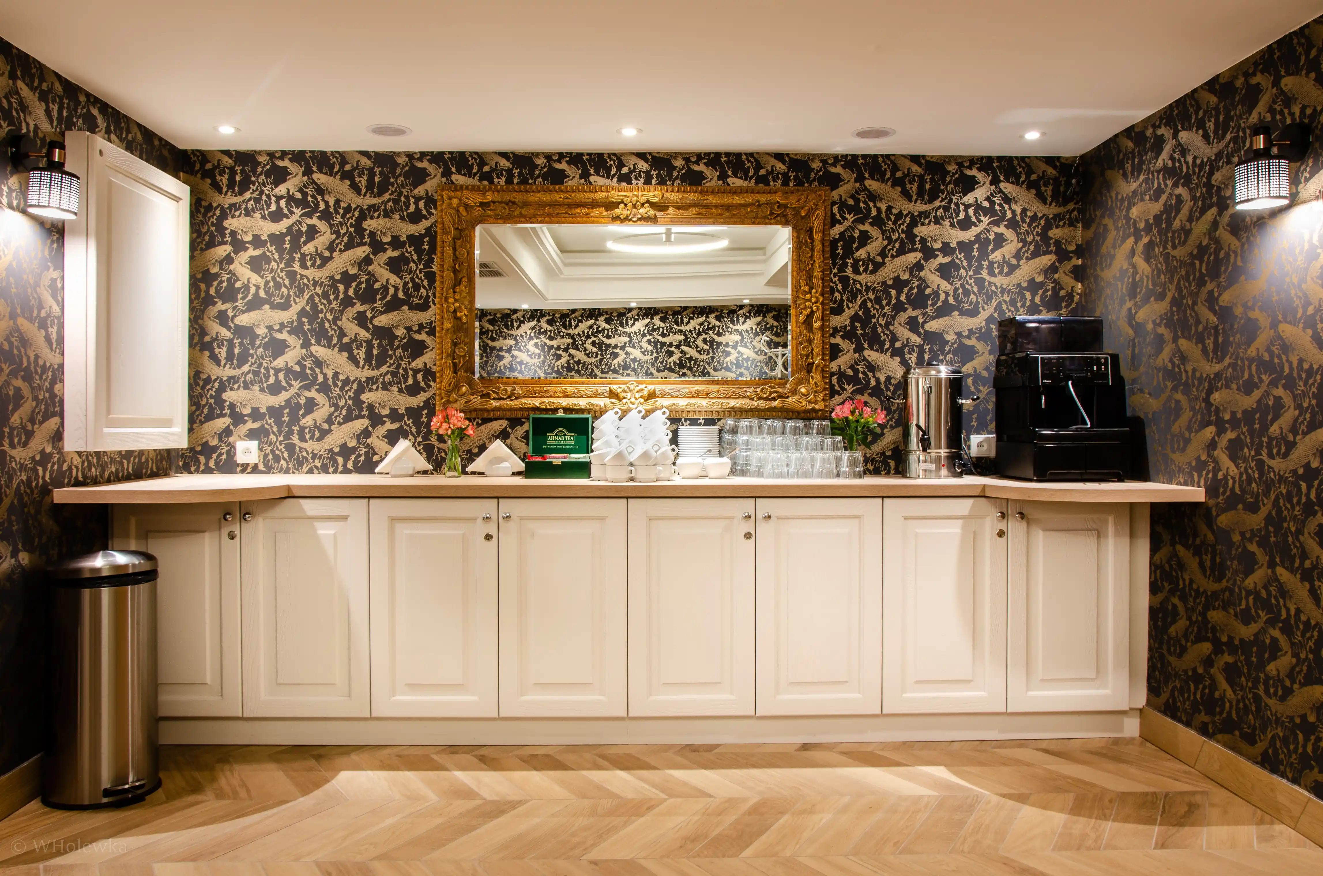 Tea and coffee station with white cabinets, ornate gold-framed mirror, floral wallpaper, and hardwood floor.