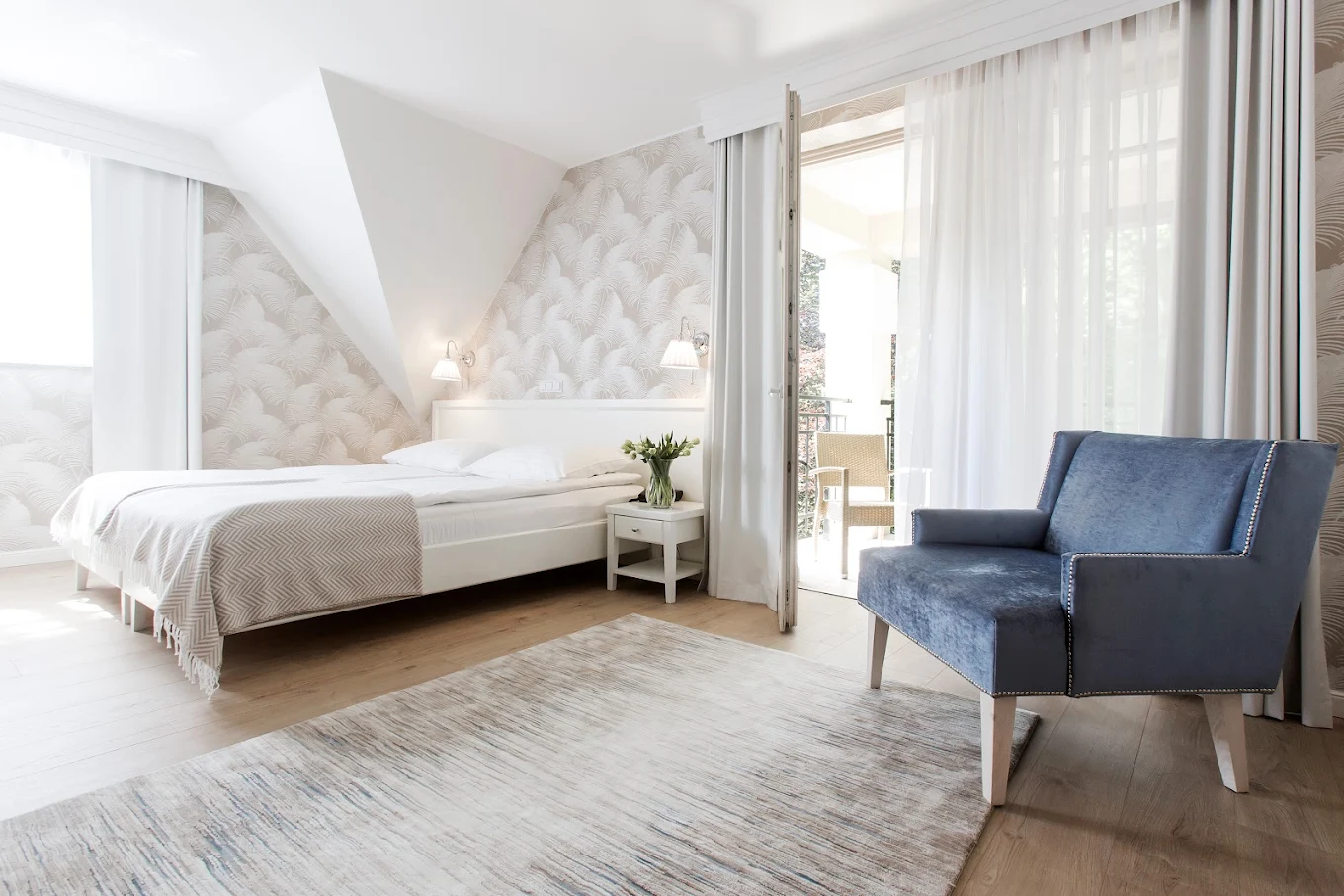 Bright bedroom with white bed, patterned wallpaper, blue armchair, and glass door leading to balcony.
