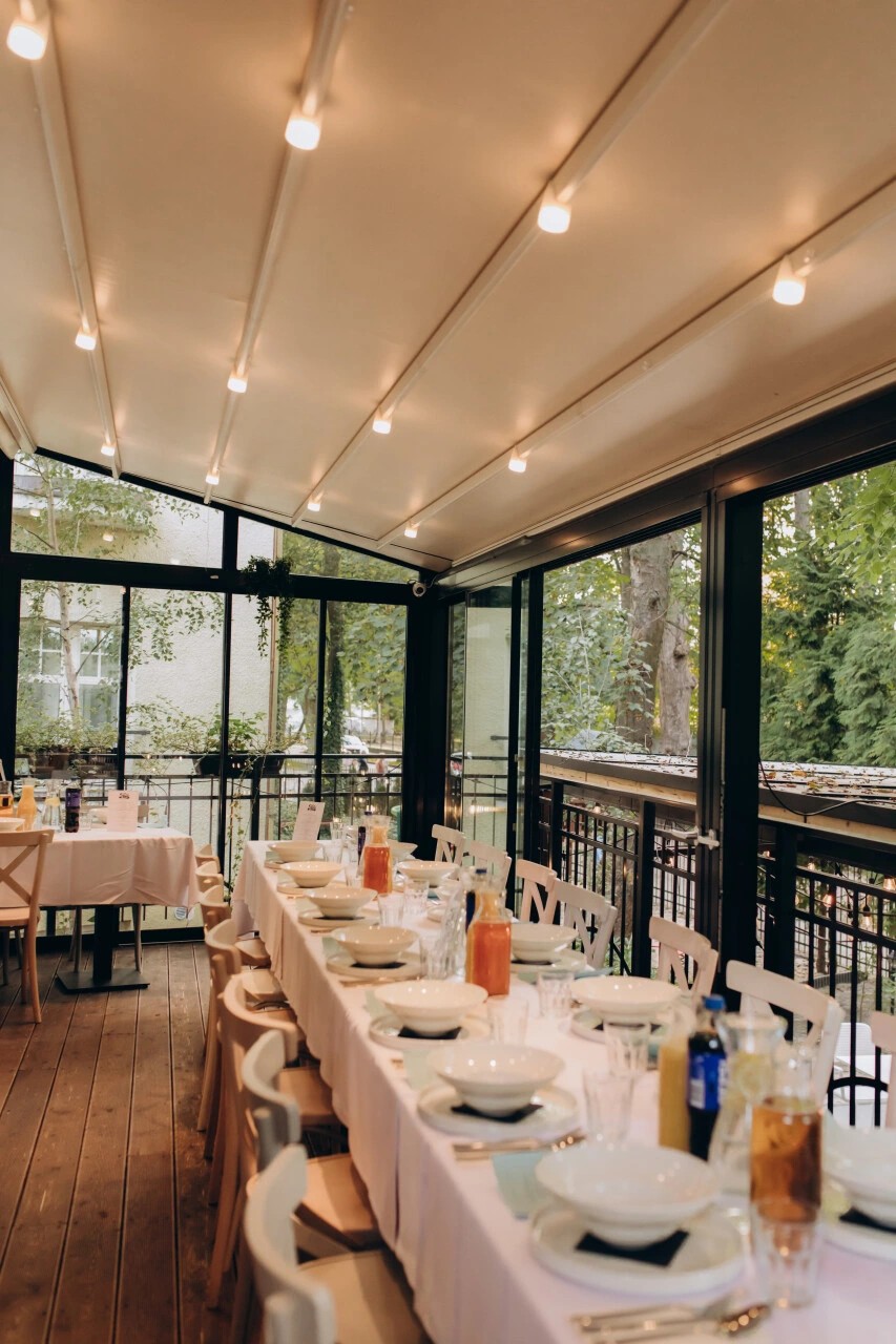 Long dining table set with white plates, bowls, glasses, and sauces under a covered outdoor patio with string lights.