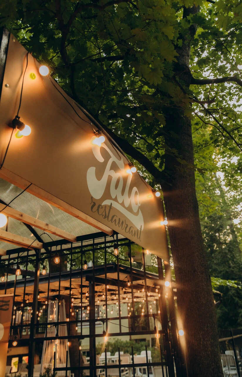 Outdoor restaurant entrance with string lights hanging under a canopy among green trees.