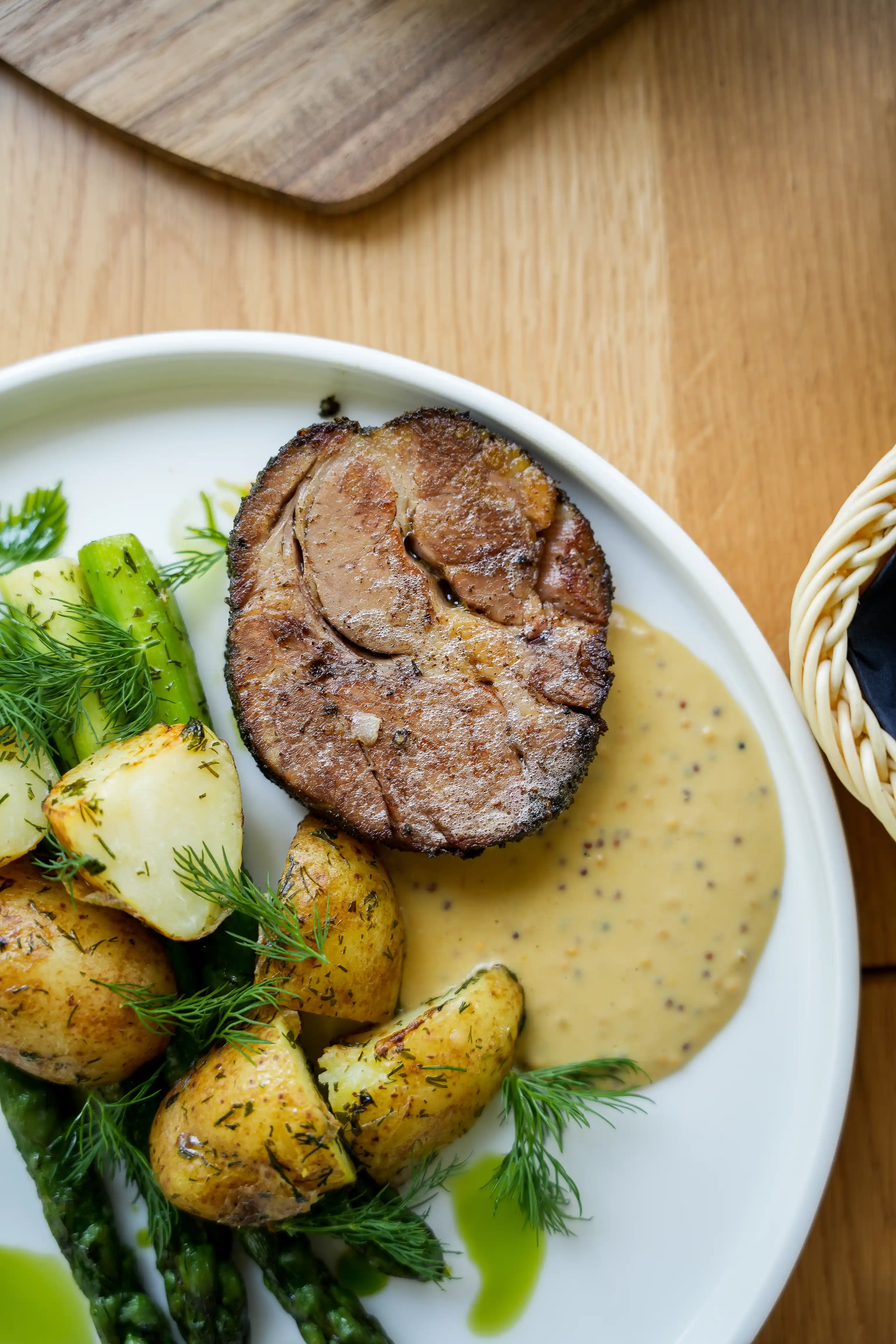 Plate with roasted potatoes, asparagus, a slice of cooked meat, fresh dill, and a creamy mustard sauce on a wooden table.