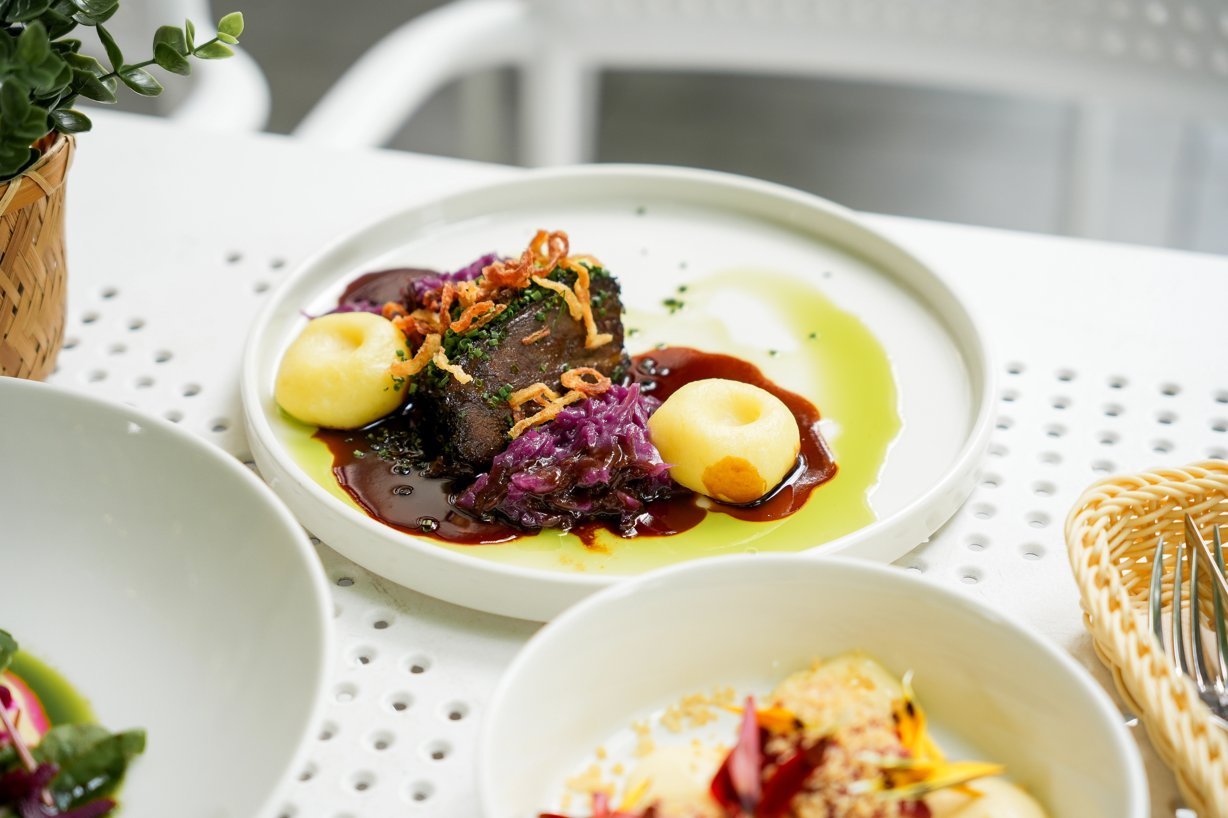 Gourmet dish with a piece of meat topped with crispy onion, surrounded by two dollops of mashed potatoes, purple cabbage, and sauces on a white plate.