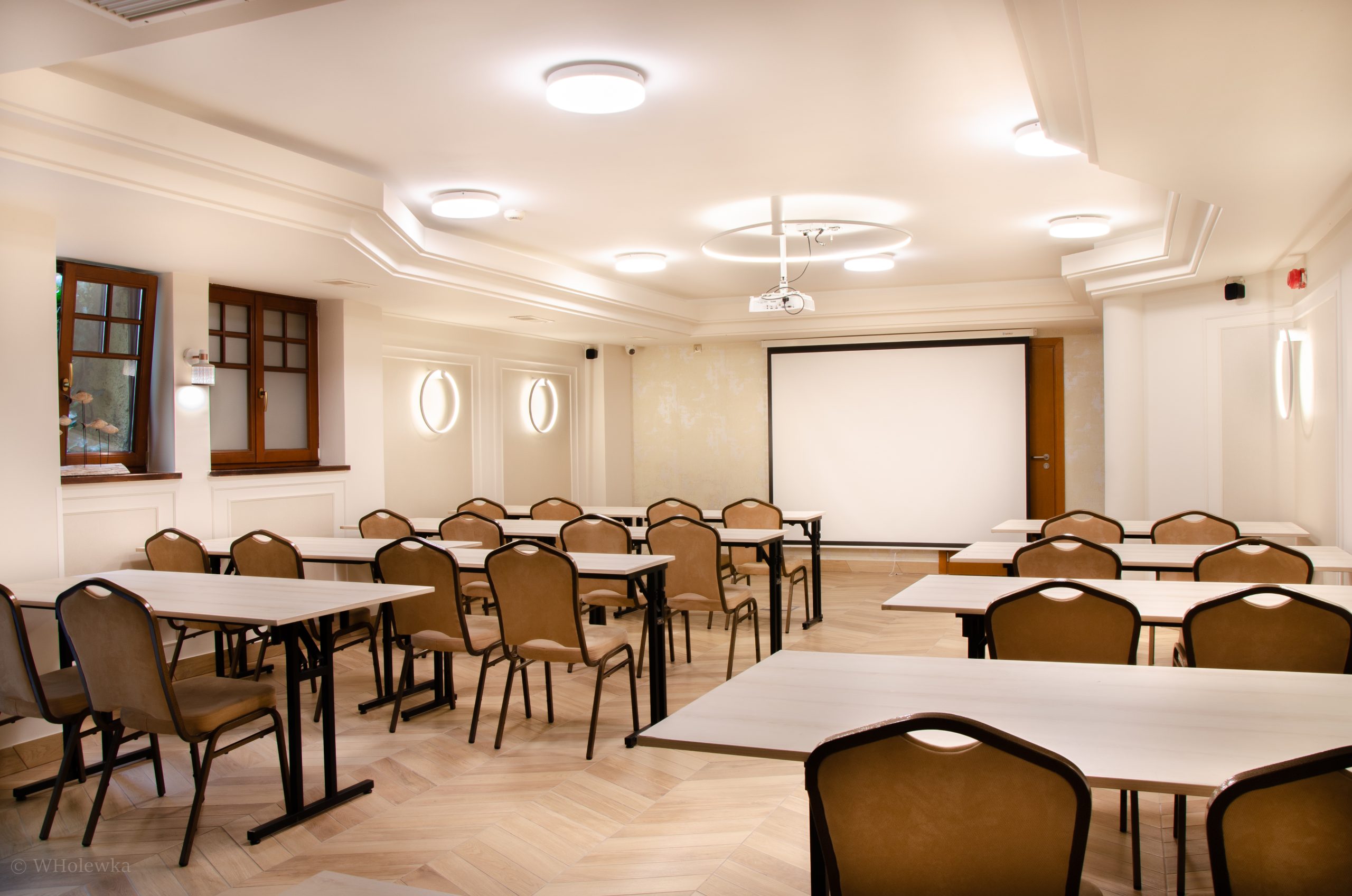Conference room with multiple rows of beige chairs and white tables facing a white projection screen on the wall.