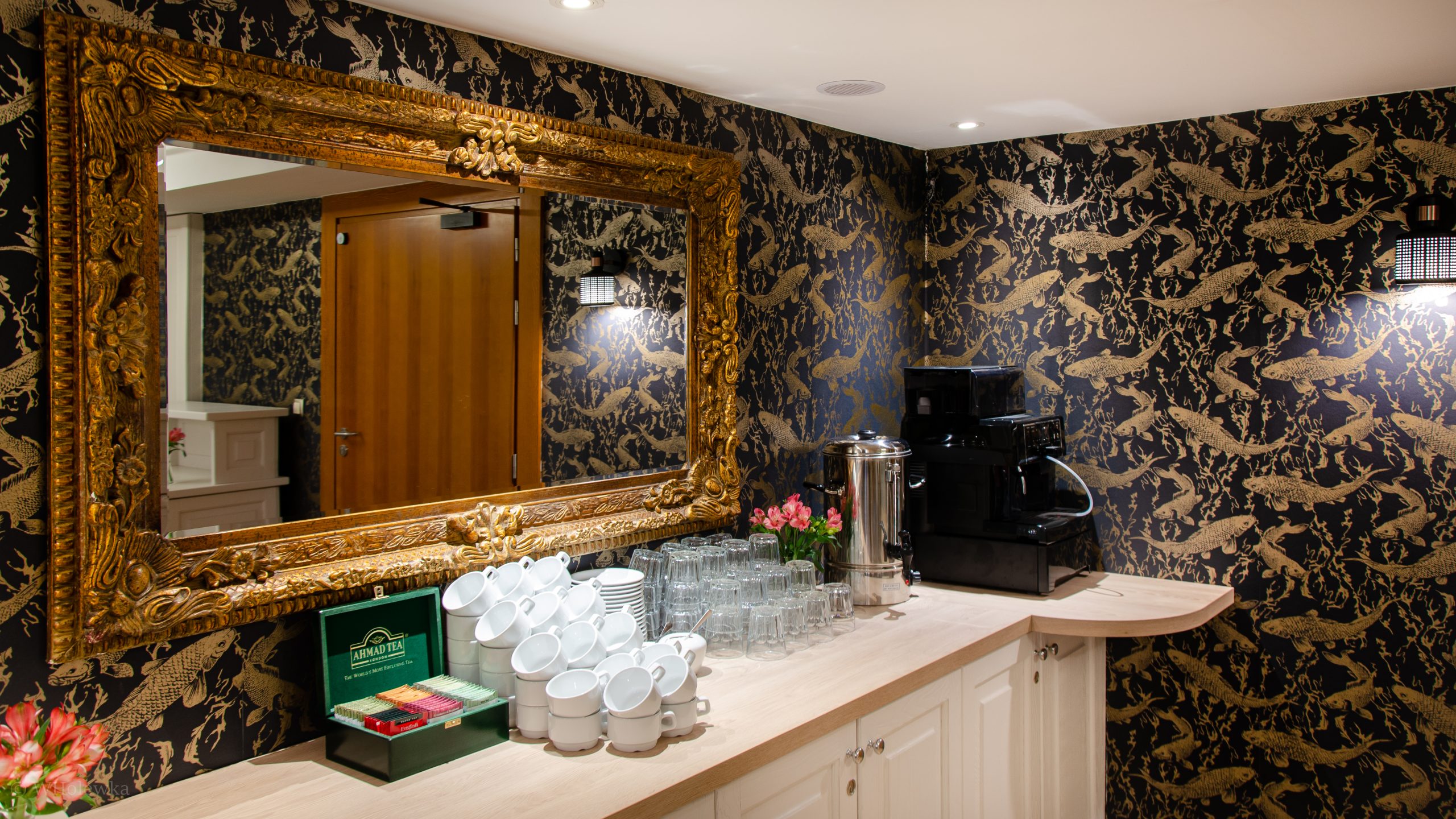A tea and coffee station with stacked white cups, clear glasses, a tea box, a silver urn, and a black coffee machine against black and gold fish-patterned wallpaper and a large ornate gold-framed mirror.