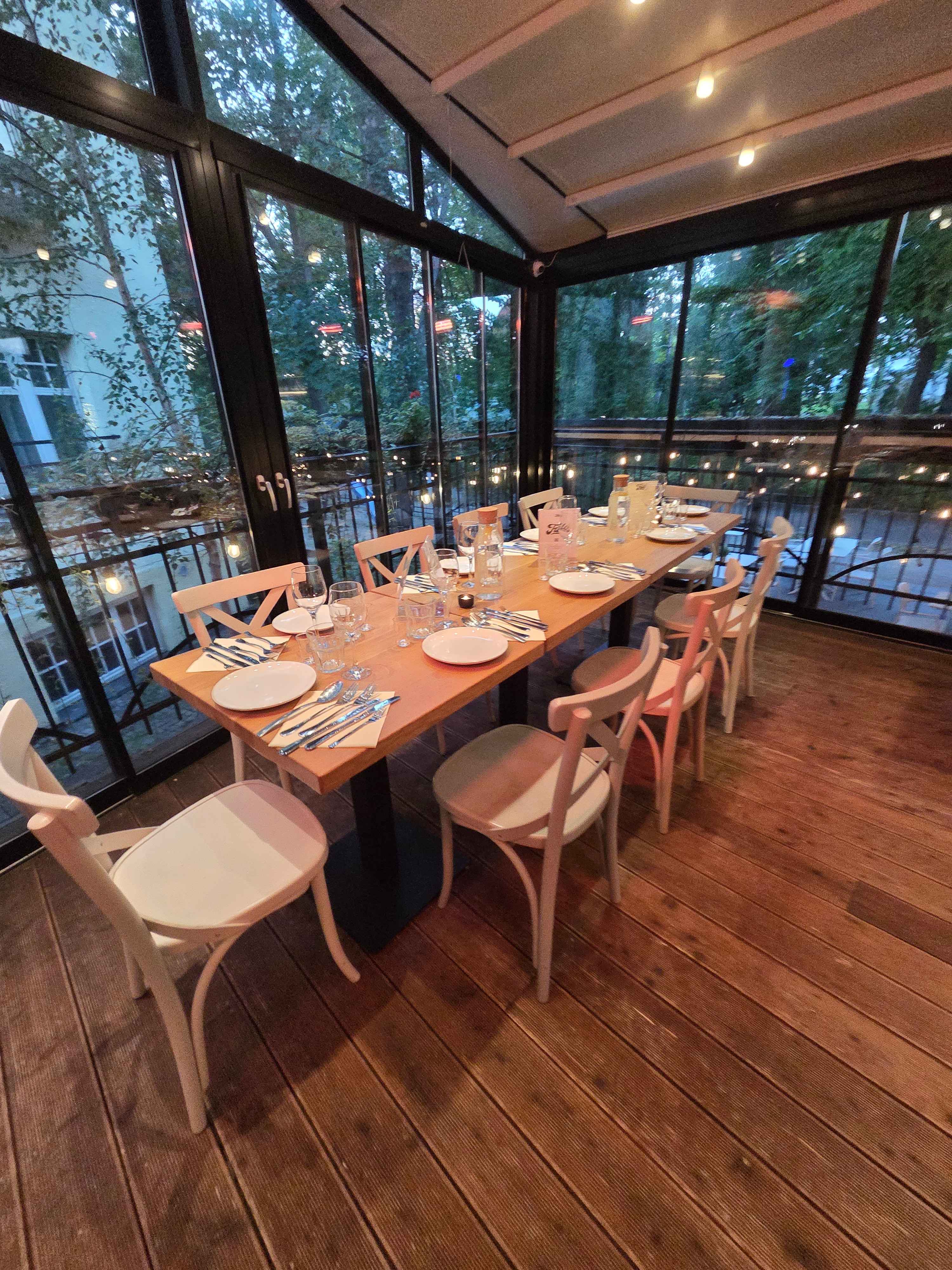 Long wooden table set for dinner with white plates, glasses, utensils, and water bottles in a glass-enclosed patio with wooden floor and trees outside.