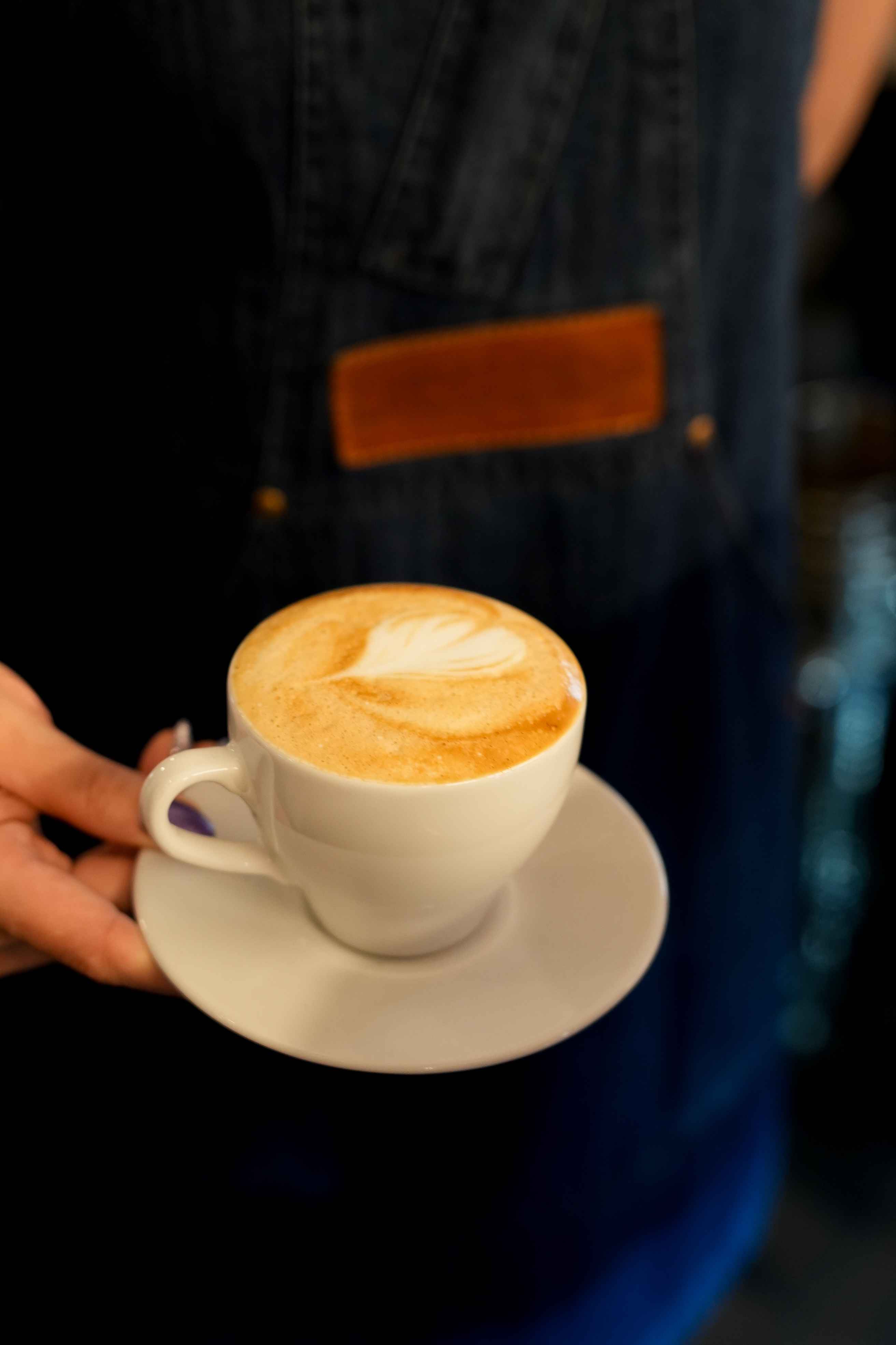 White cup of cappuccino with heart-shaped latte art on a white saucer held by a person wearing denim overalls.