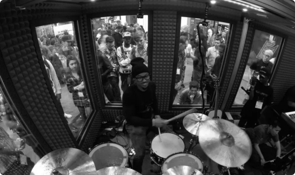 Drummer inside a large drum booth at the NAMM show, with crowds of people watching through the windows of the double-walled soundproof booth.