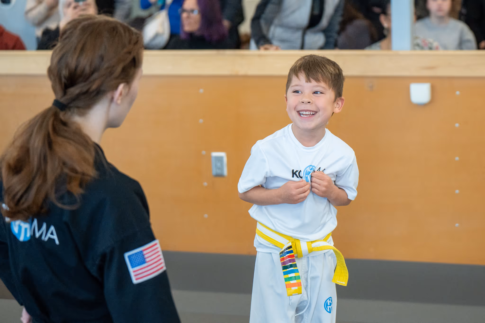 Smiling young boy with a yellow martial arts belt facing a woman with an American flag patch on her black uniform.