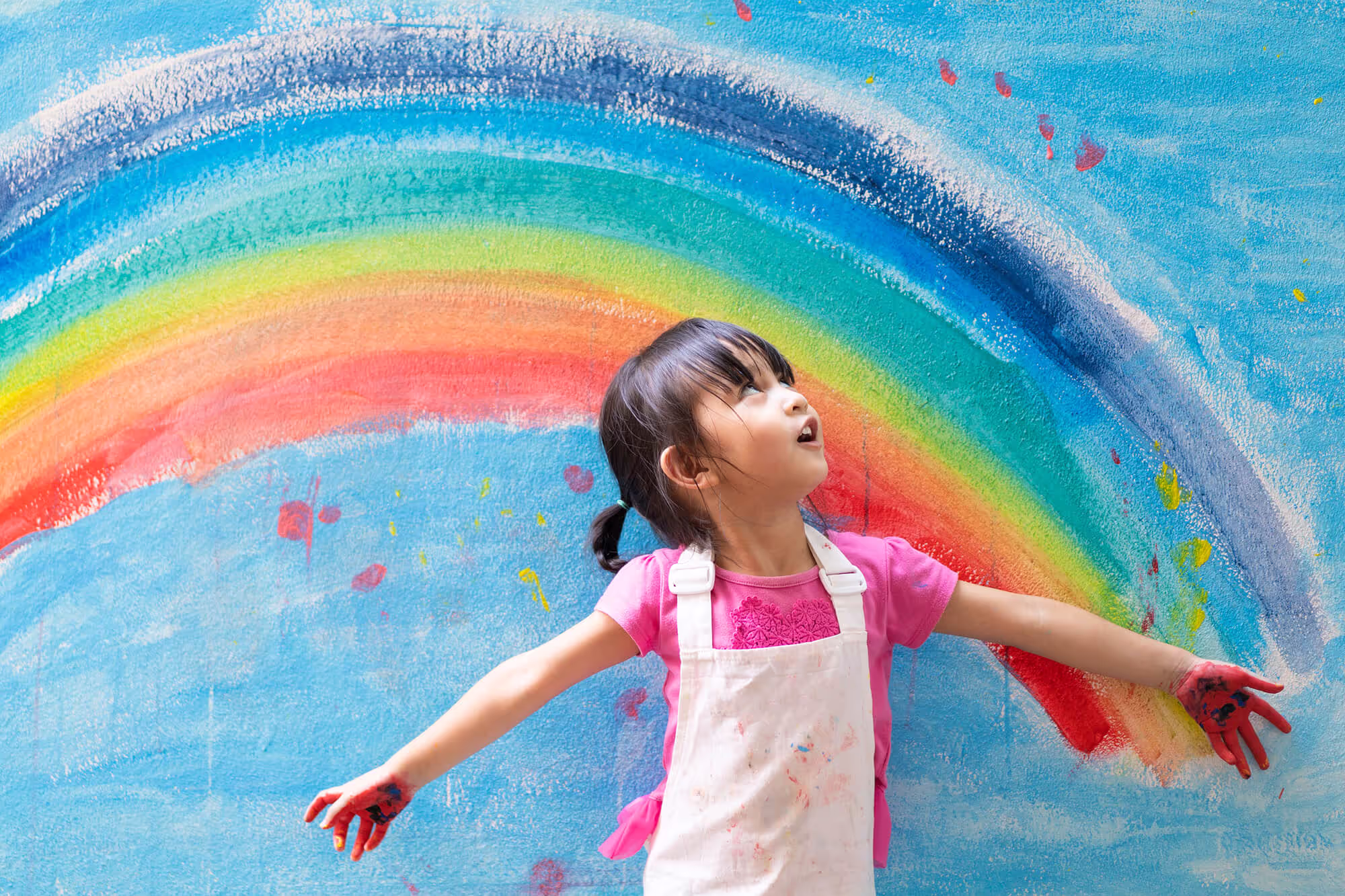 A little girl standing in front of a rainbow painted wall.