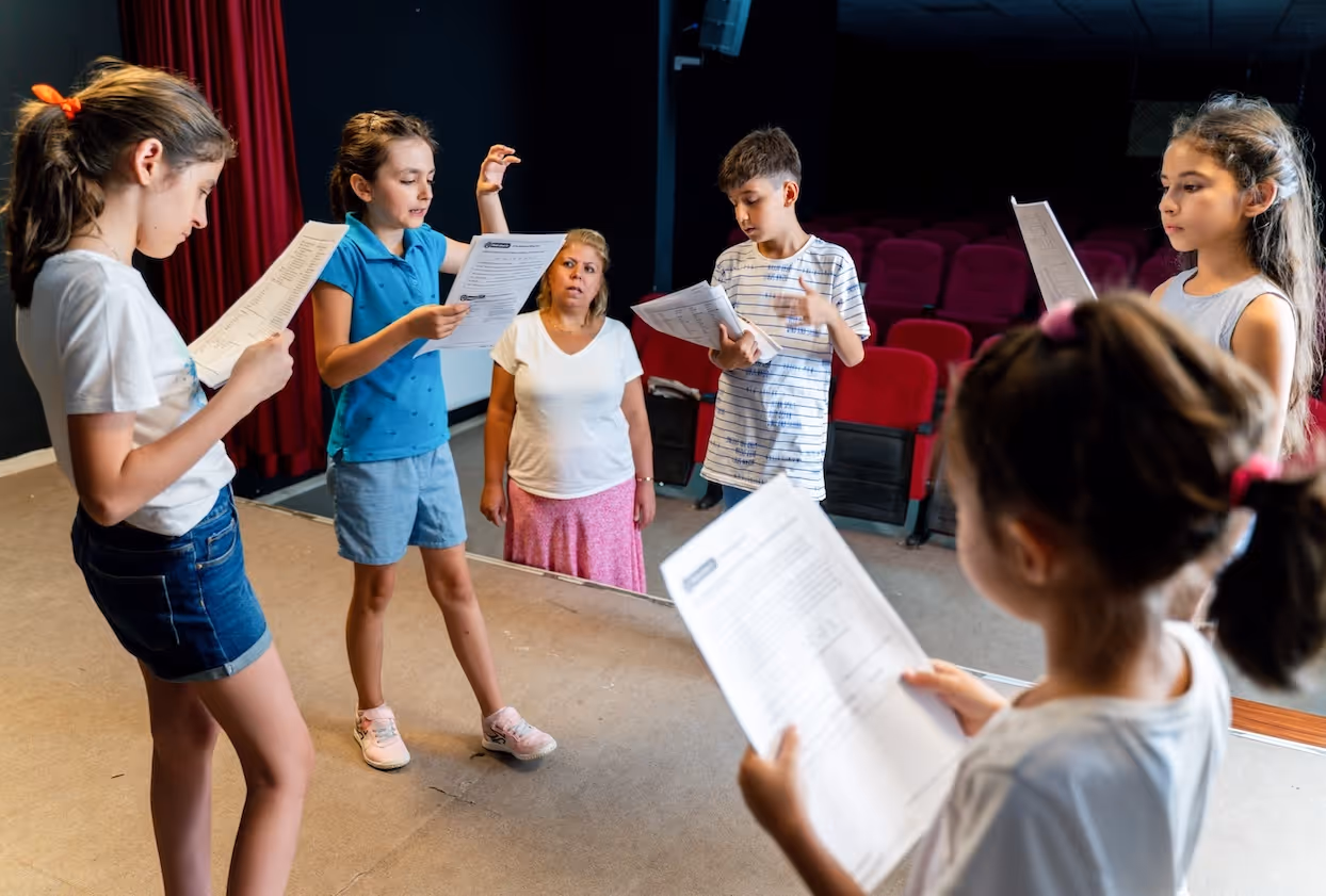 A group of kids stand on a stage in a circle holding scripts. An instructor watches from off stage.