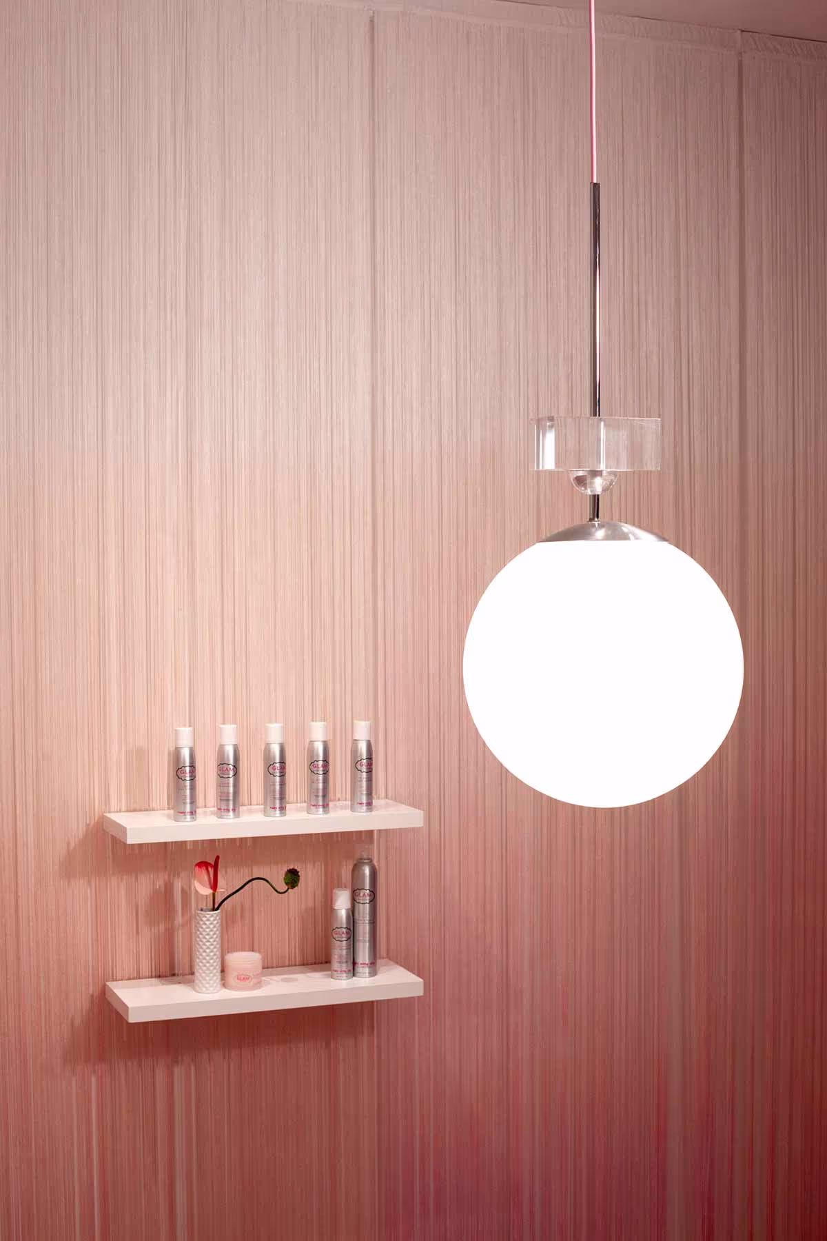 White shelving with curated product display against pink backdrop and oversized custom globe light. Beauty product merchandising in luxury salon interiors.