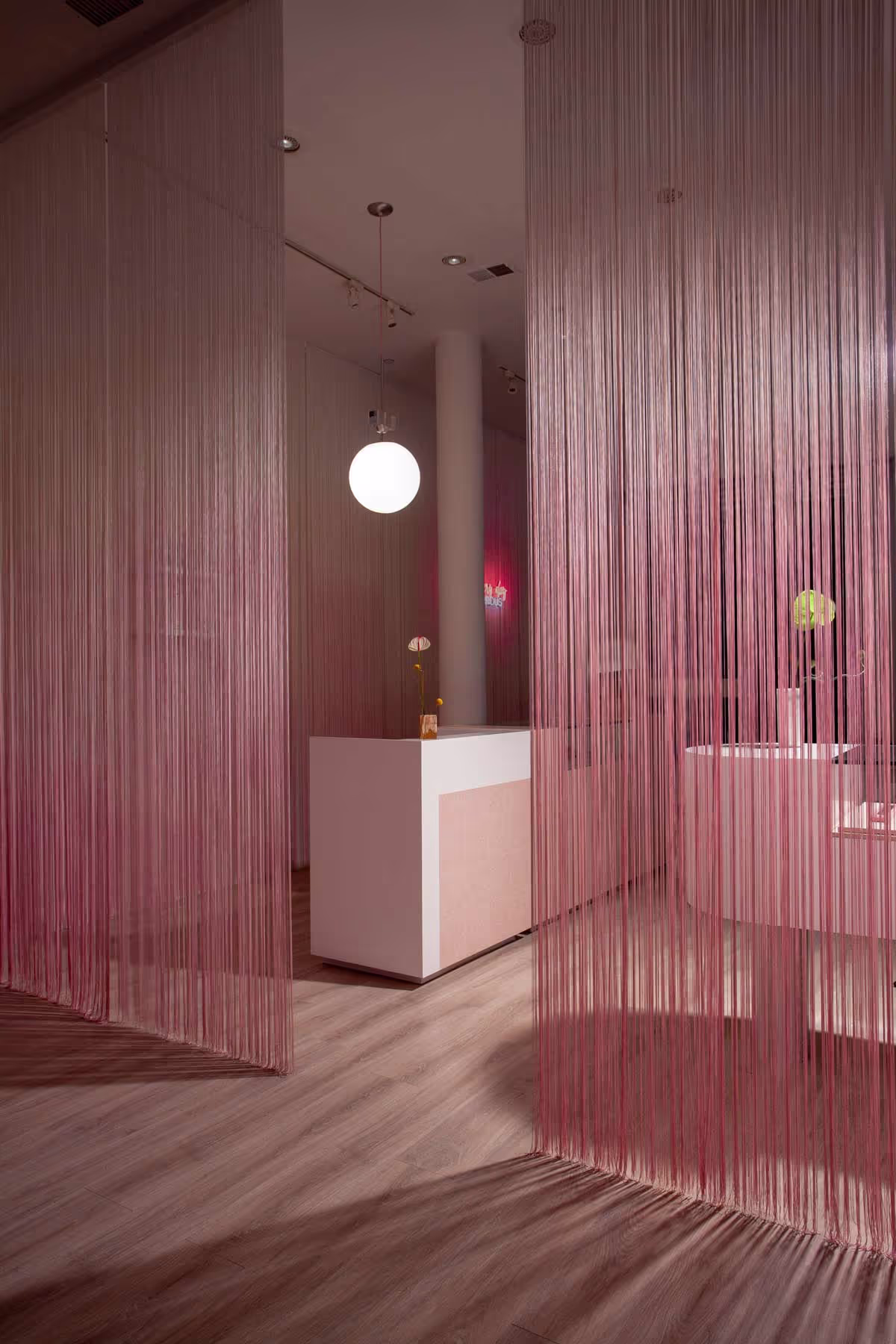 Salon reception with ombré pink thread dividers and floating counter. Retail-focused hair salon design with sculptural elements and soft textures.