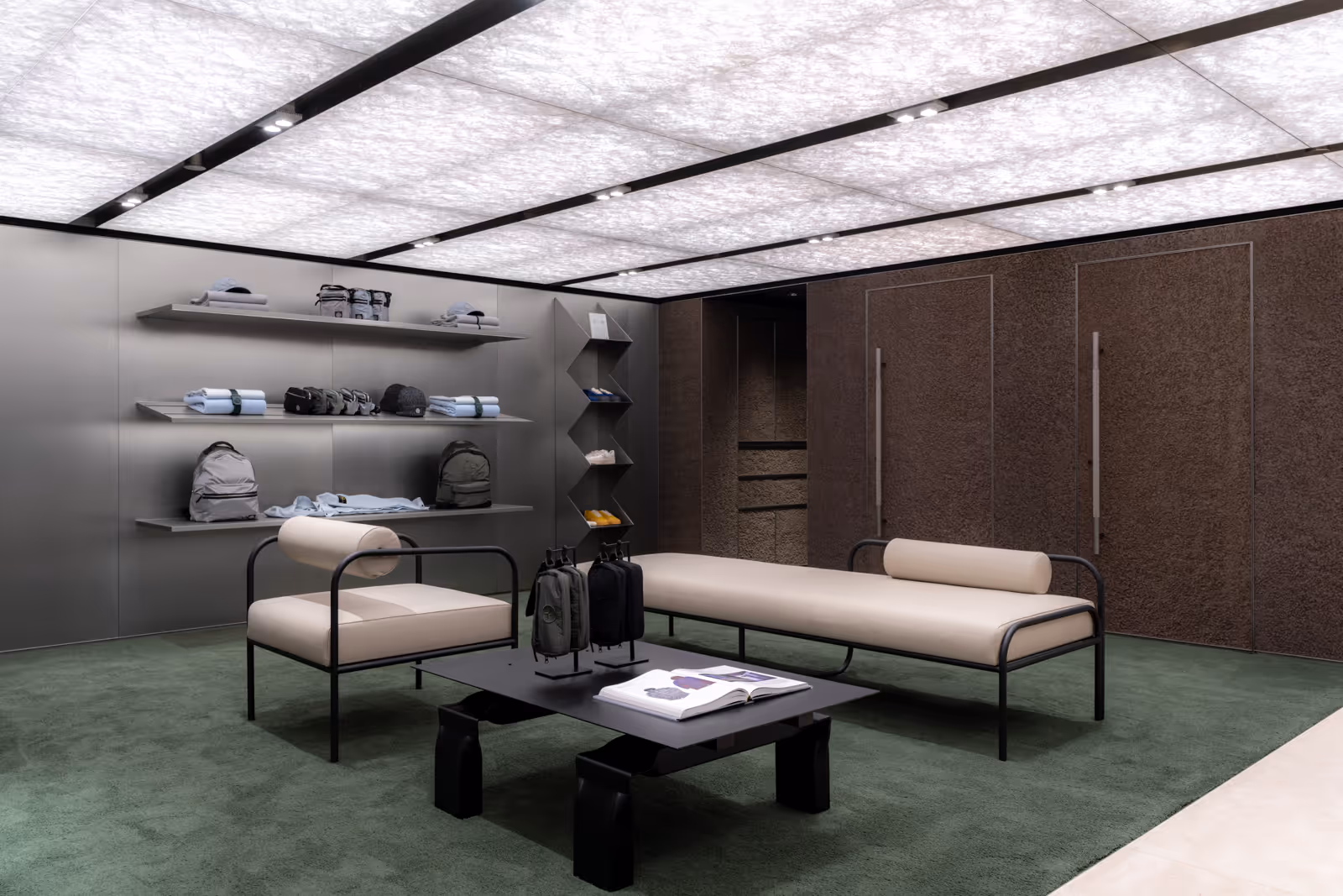 Modern Stone Island flagship store interior in Hong Kong featuring custom-designed beige cushioned daybed and armchair by Sergio Mannino Studio with black matte metal frames, minimalist shelving, green carpet, and soft grid ceiling lighting.