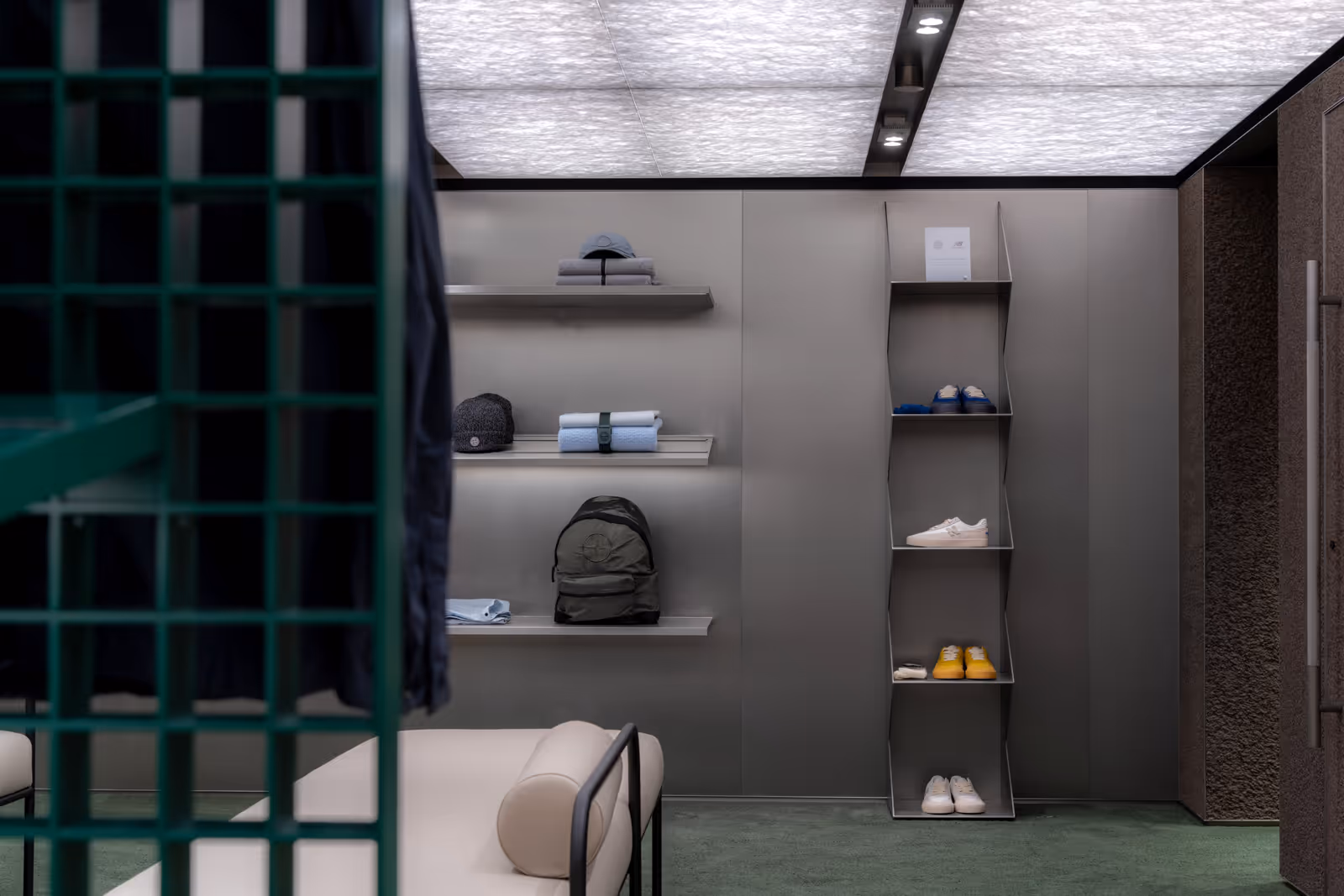 Display wall inside Stone Island Hong Kong flagship store with view of the Shiro daybed, accessories, and shoes on metallic shelves, with a green metal grid structure, recessed ceiling lighting, and green carpet flooring.