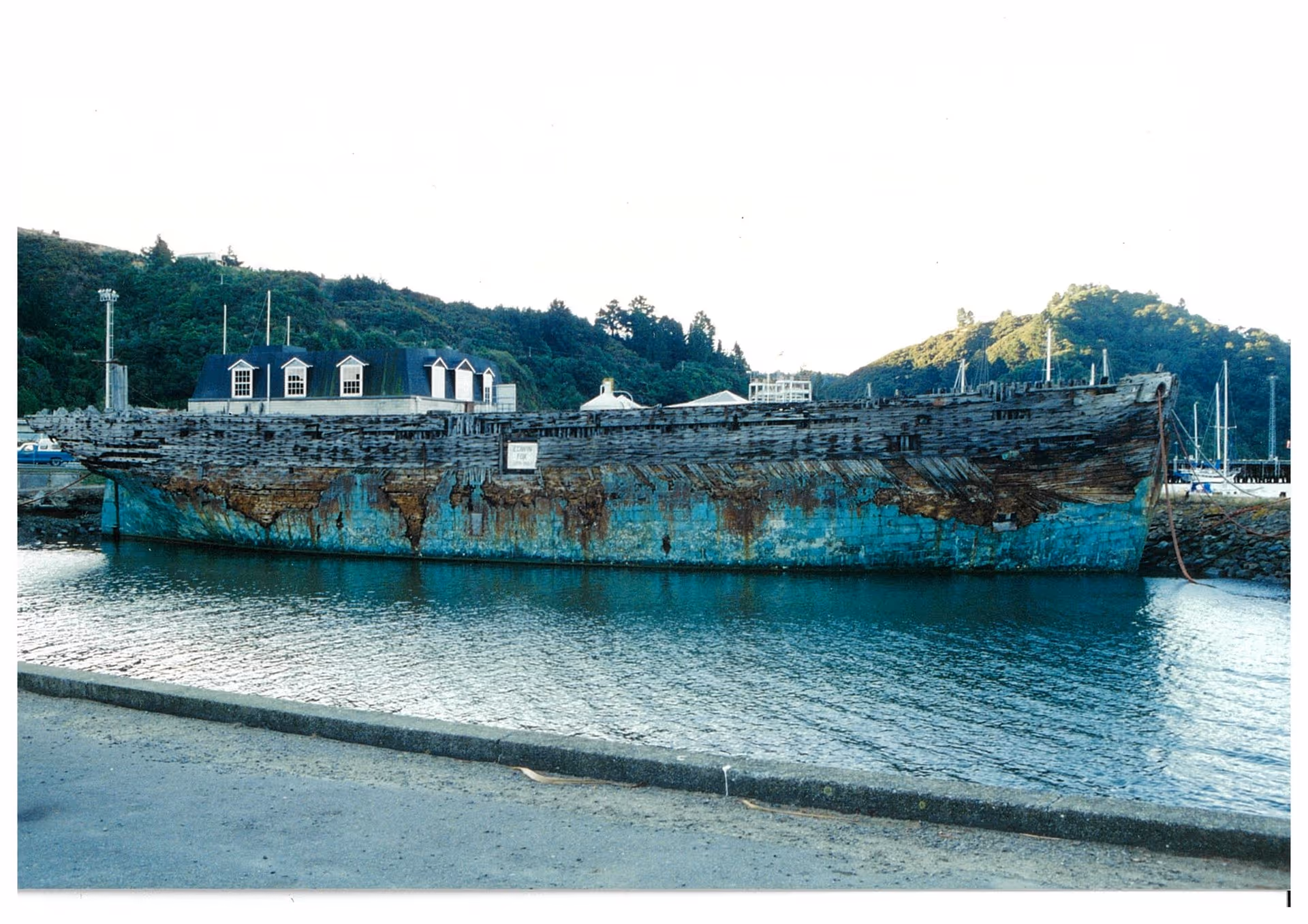 An old, weathered ship with peeling blue paint rests in a calm harbor. Small buildings are on deck, surrounded by lush, green hills in the background.