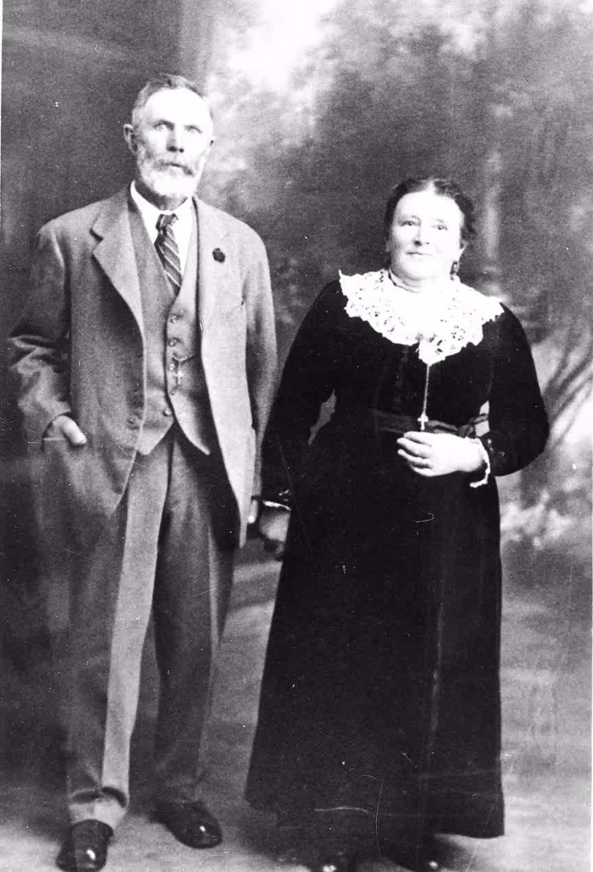 An older man in a suit and tie stands beside a woman in a long dark dress with a lace collar, posing for a formal studio portrait.