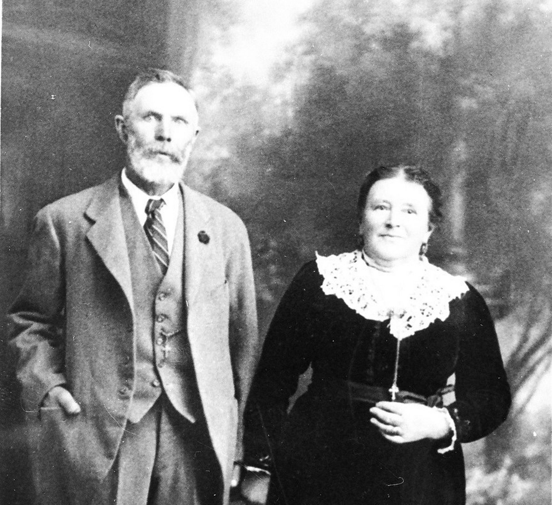 An older man in a suit and tie stands beside a woman in a long dark dress with a lace collar, posing for a formal studio portrait.