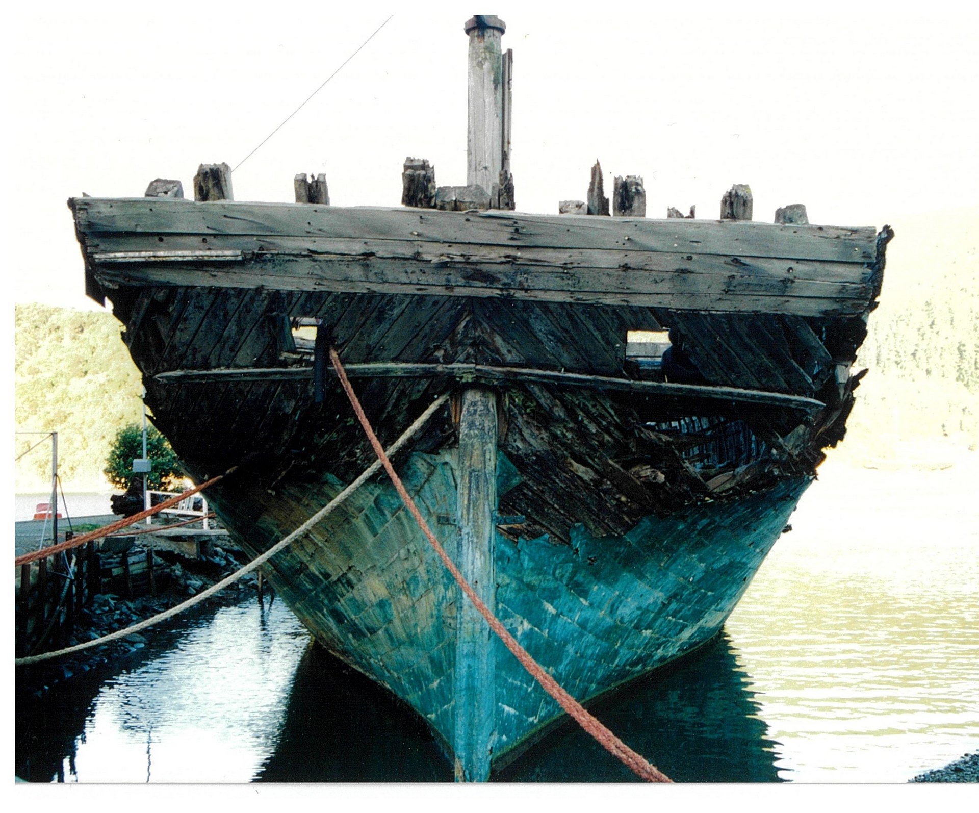A large, weathered wooden ship with a deteriorating hull is moored at a dock, viewed from the front.