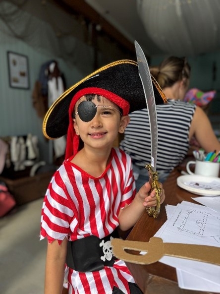 A child dressed as a pirate in a striped red and white costume, wearing a large hat and holding a toy sword, sits at a table.