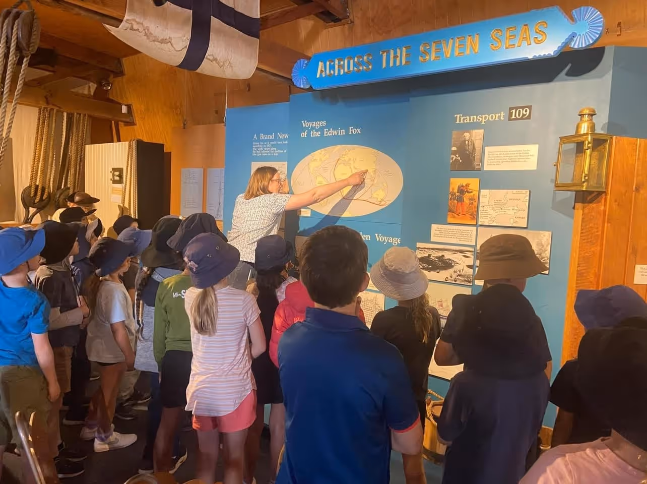 A teacher points to a world map while engaging a group of children wearing hats in a maritime exhibit about the Edwin Fox.