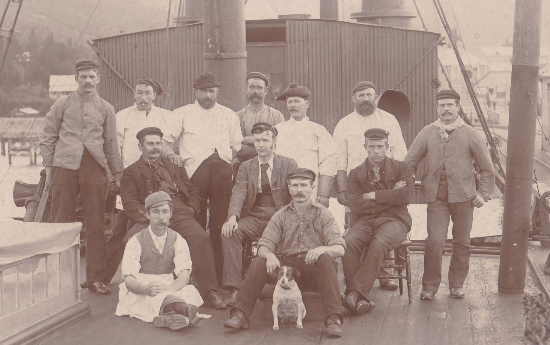 A group of twelve men in period attire, posing on a wooden deck of a boat, with a small dog seated in front of them.