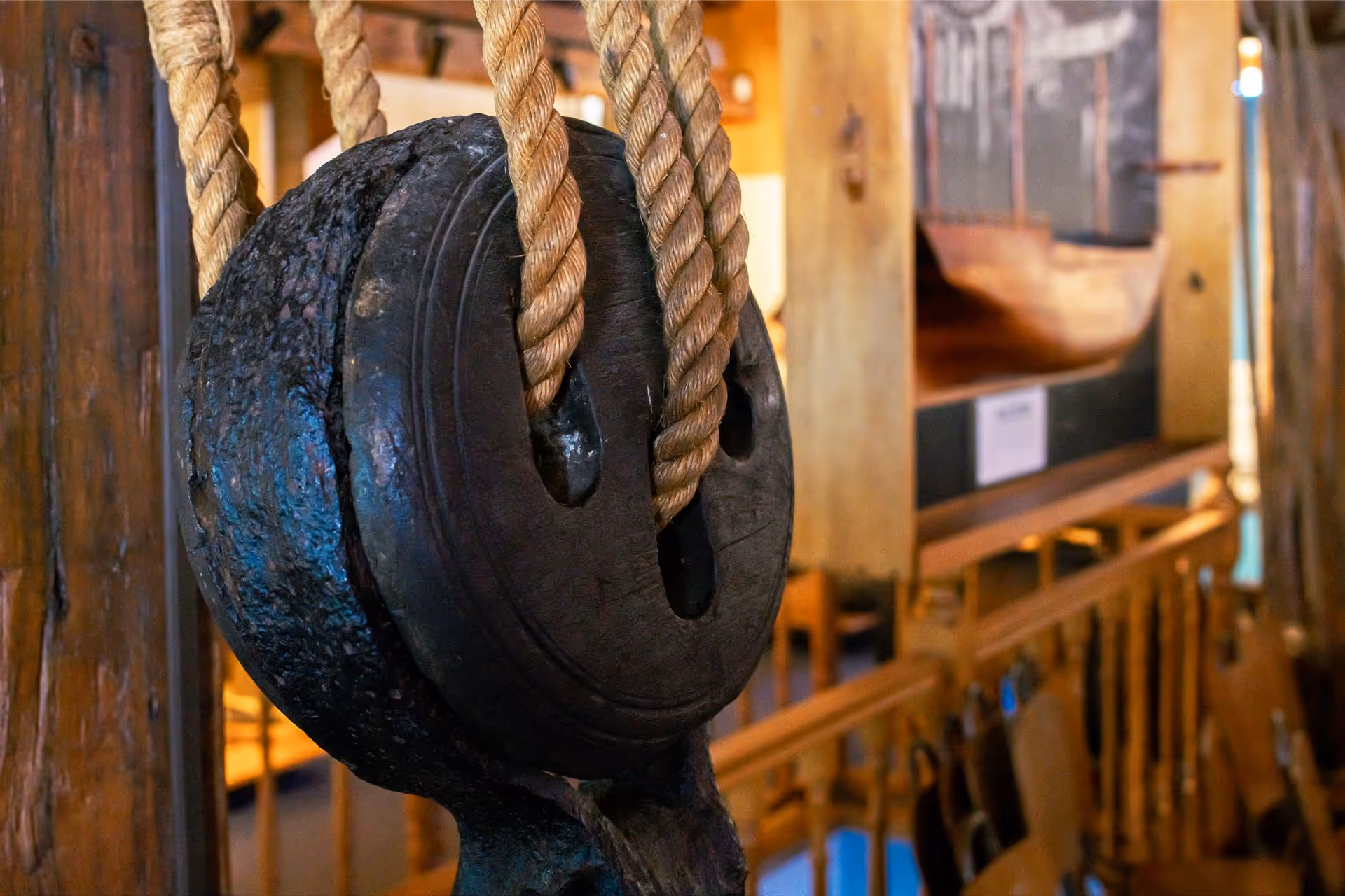 Close-up of a weathered wooden pulley with thick ropes, set against a blurred maritime background of a museum exhibit.