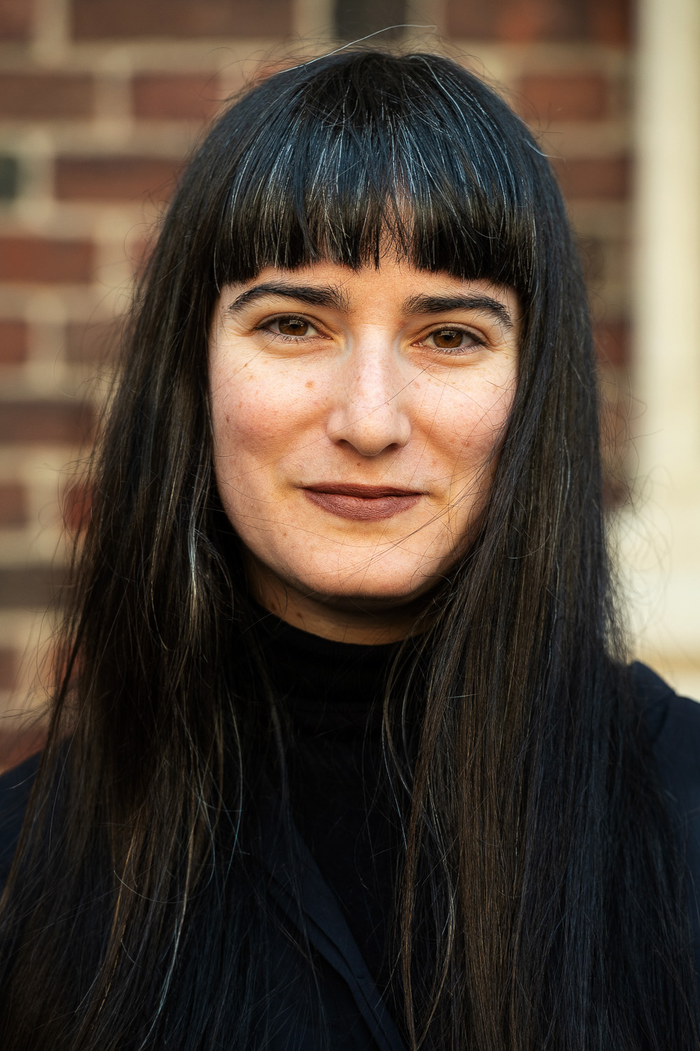 Woman with bangs standings looking at camera with brick wall in background. 