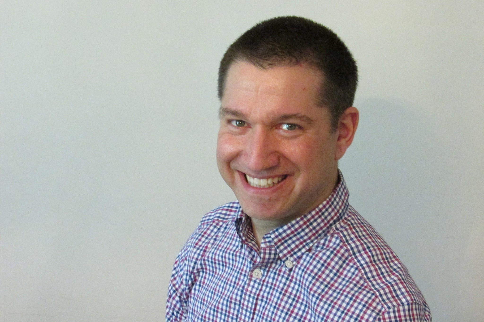 Man with plaid shirt smiling at camera with blank wall behind him. 