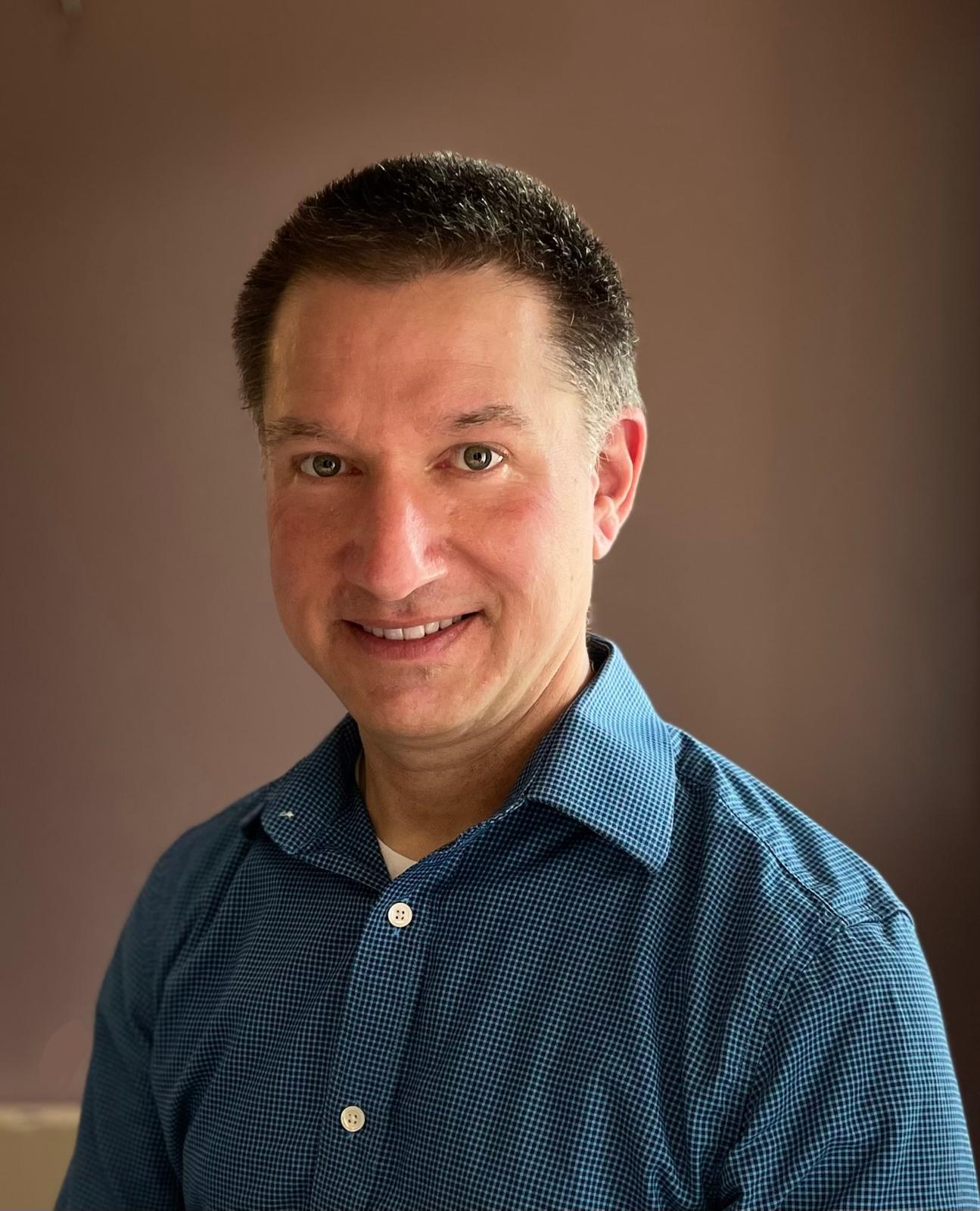 Man with plaid shirt smiling at camera with blank wall behind him. 