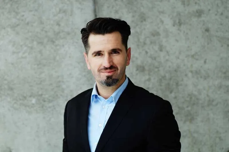 Porträt von Lulzim Zhuta von Forward Energie, Mann im schwarzen Sakko vor Betonwand.