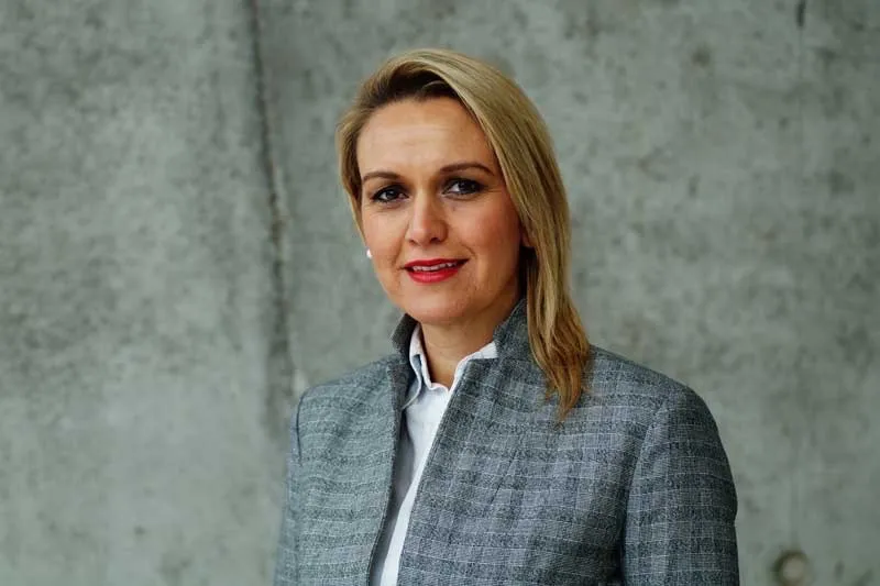 Porträt von Suzana Zhuta von Forward Energie, Frau im grauen Blazer vor Betonwand.