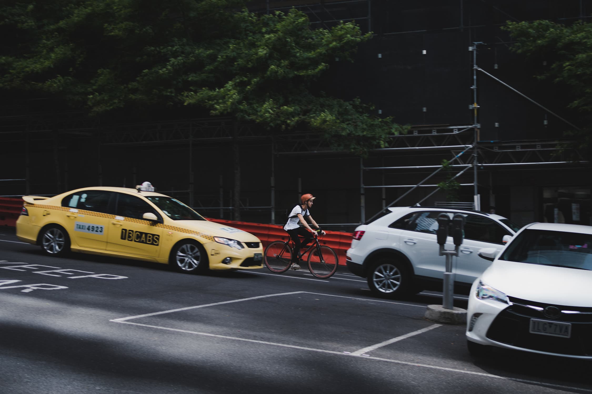 Person wearing orange helmet riding a bike between yellow taxi and white parked cars on city street.